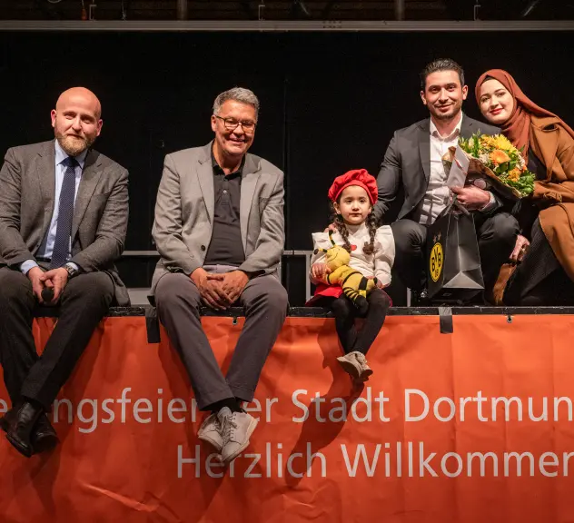 Fünf Personen sitzen auf dem Rand einer Bühne, auf einem Transparent vorne an der Bühne steht "Einbürgerungsfeier der Stadt Dortmund. Herzlich Willkommen"