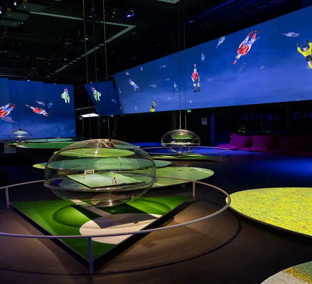 Ein Raum mit Glaskugeln, in denen Miniatur-Fußballfelder stecken. An die Wände sind Bilder von Kicker-Figuren projiziert.