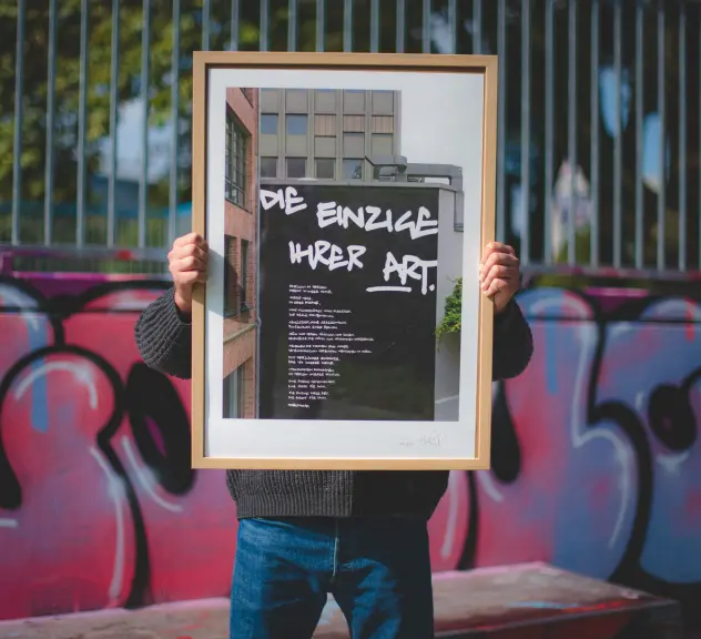 Mann steht vor einer bunten Graffiti-Wand und hält ein Bild mit Holzrahmen hoch. Das Bild ist ein Foto eines Wandgemäldes in Dortmund