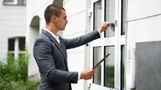 Mann im Anzug und mit Klemmbrett in der Hand klopft an eine Haustür