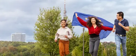 Drei Jugendliche rennen. Im Hintergrund ist der Florianturm zu sehen. Das Mädchen in der Mitte lässt eine Europa-Flagge hinter sich flattern, alle drei lachen. 