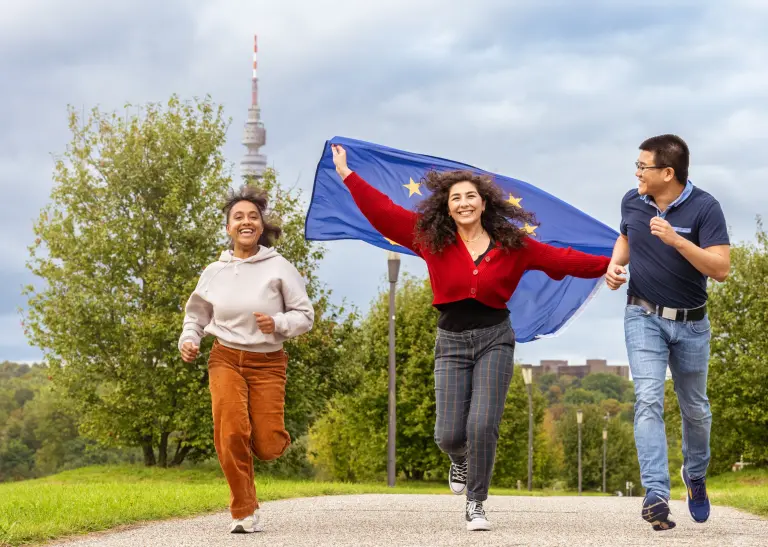 Drei Jugendliche rennen. Im Hintergrund ist der Florianturm zu sehen. Das Mädchen in der Mitte lässt eine Europa-Flagge hinter sich flattern, alle drei lachen. 