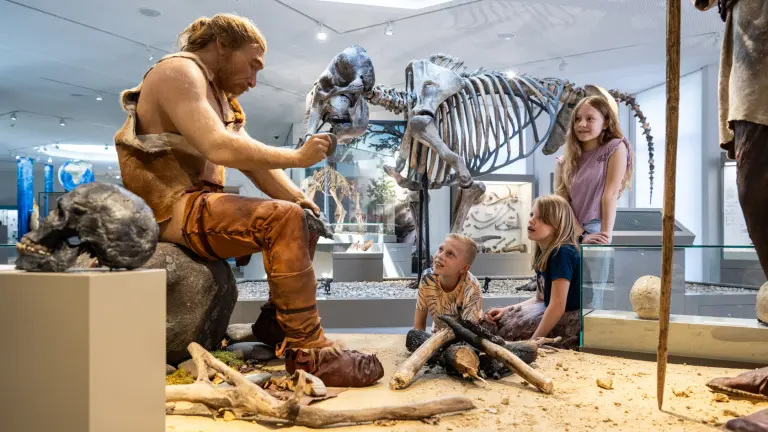 Ein großer Mann, verkleidet als Neandertaler, sitzt auf einem Stein und zeigt vier staunenden Kindern, wie er Feuer mit einem Stein macht