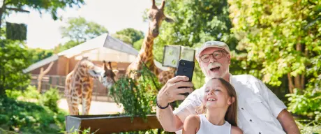 Ein älterer Mann und ein Kind machen zusammen ein Selfie vor dem Giraffengehege des Dortmunder Zoos.