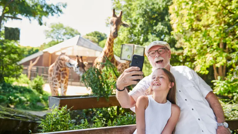 Ein älterer Herr und ein Mädchen machen lachend ein Selfie vor dem Giraffengehege im Zoo.