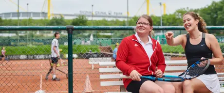 Zwei Frauen sitzen mit Tennisschlägern in der Hand auf einer Bank. Im Hintergrund sieht man einen Tennisplatz auf dem Tennis gespielt wird und den Signal Iduna Park