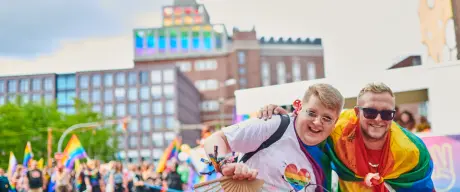 Zwei Personen lachen auf einer LGBTQIA-Veranstaltung in die Kamera. Im Hintergrund zeigt der Dortmunder U-Turm die LGBTQIA-Flagge.