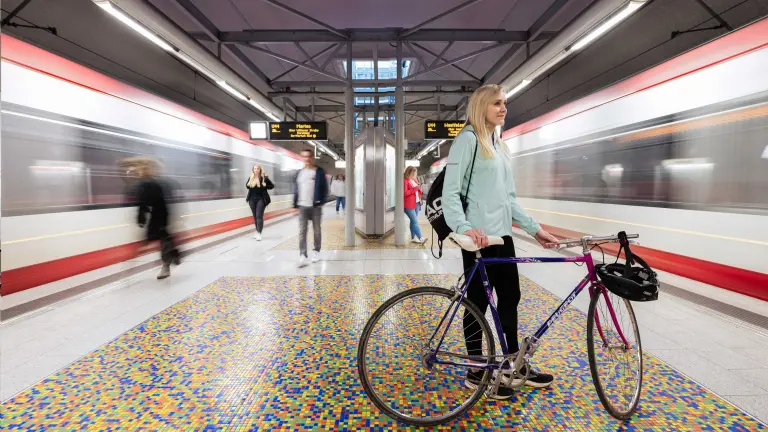Man sieht eine U-Bahnstation in Dortmund, zwei Bahnen fahren vorbei, im Vordergrund steht eine Frau mit Ihrem Fahrrad, im Hintergrund sind weitere Personen zu sehen.