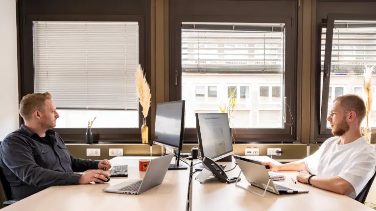 Zwei Personen sitzen gegeneinander im Büro und arbeiten jeweils an ihren Schreibtischen
