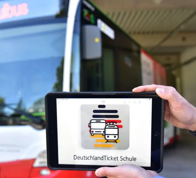 Im Vordergrund ist ein Tablet, dass von zwei Händen gehalten wird. Auf dem Display steht Deutschland Ticket Schule. Im Hintergrund steht ein Bus, auf der Anzeige steht "Schulbus".