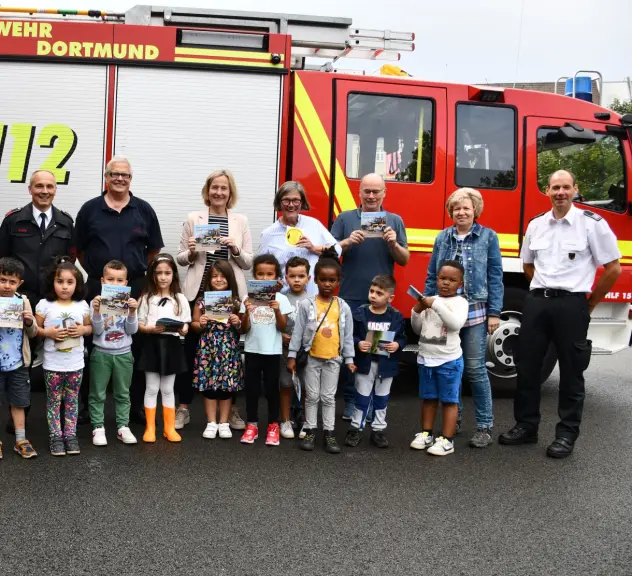 Vertreter*innen der Feuerwehr Dortmund, der Fabido-Kita Bornstraße und der Stadt Dortmund stehen vor einem Feuerwehrauto, in der vordersten Reihe stehen außerdem zehn Kinder aus der Fabido-Kita. Alle halten das neue Plappermaul-Buch hoch.