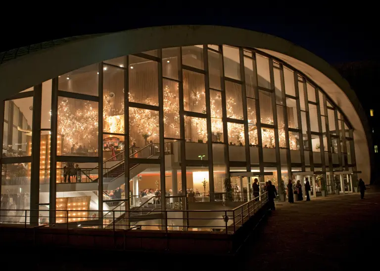 Das beleuchtete Opernhaus Dortmund bei Nacht, drinen hinter der Glasfassade und draußen stehen Menschen.