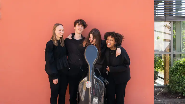 Vier junge Menschen stehen vor einer orangenen Wand. In der Mitte ist ein Cello-Koffer. 
