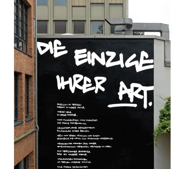 Kunstdruck "Die einzige Ihrer Art" auf einer großen Wand in einem Innenhof. Zu lesen ist das Markennarrativ in weißer Graffiti-Schrift auf schwarzem Grund. 