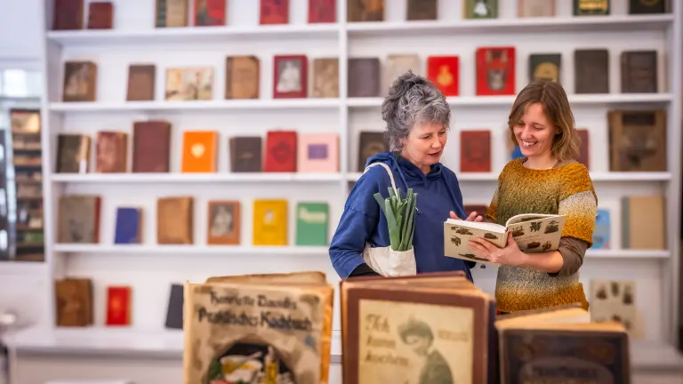 Zwei Frauen stehen im deutschen Kochbuchmuseum und schauen sich lächelnd gemeinsam ein Buch an, während aus der Handtasche einer der Frauen ein paar Stangen Lauch herausschauen