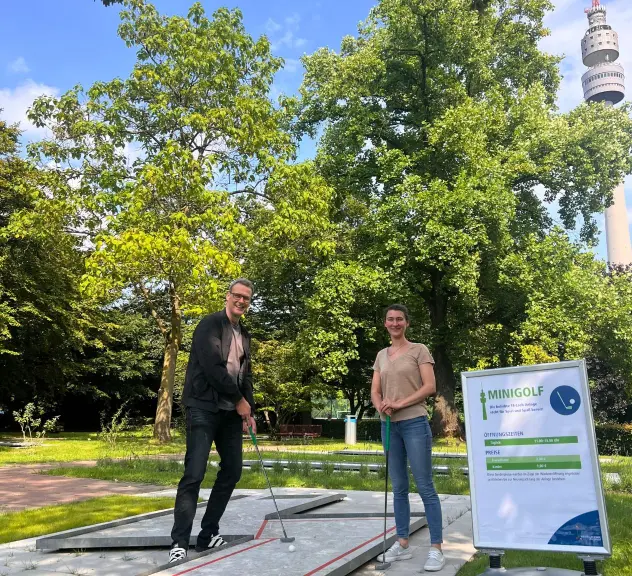 Zwei Personen spielen Minigolf im Westfalenpark Dortmund, umgeben von grünen Bäumen und dem Fernsehturm im Hintergrund.