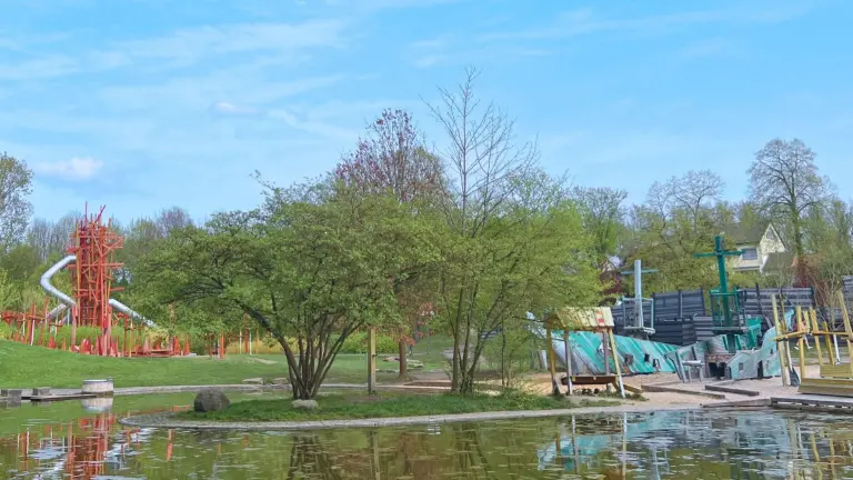  Ein farbenfroher Spielplatz und ein einzigartiger Spielbogen im Westfalenpark Dortmund, umgeben von Bäumen und einem Teich im Vordergrund. Perfekt für Kinder und familienfreundliche Ausflüge.