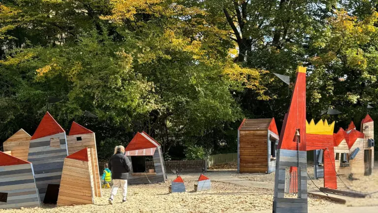 Langnese-Spielplatz im Westfalenpark Dortmund mit bunten, fantasievollen Holzhäusern und Spielstrukturen. Eine erwachsene Person und ein Kind erkunden gemeinsam die Spielgeräte, umgeben von herbstlich gefärbten Bäumen.