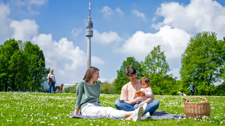 Familie genießt ein Picknick auf einer blühenden Wiese im Westfalenpark Dortmund, mit dem Florianturm im Hintergrund an einem sonnigen Tag.