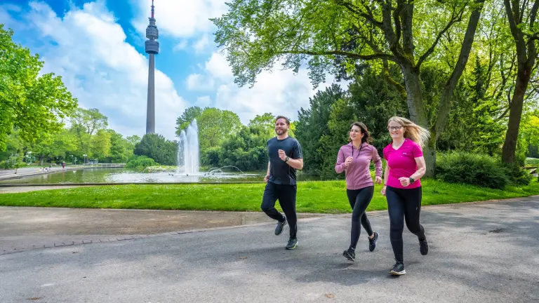 Drei Personen joggen auf einer Wegstrecke im Westfalenpark Dortmund, vorbei an einem Teich mit Springbrunnen und dem Florianturm im Hintergrund.