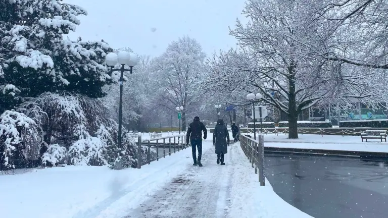 Verschneiter Westfalenpark Dortmund im Winter, mit schneebedeckten Bäumen, Laternen und einem Weg, auf dem Spaziergänger*innen unterwegs sind. Der angrenzende Teich ist teilweise zugefroren und von Bänken und Bäumen umrahmt.