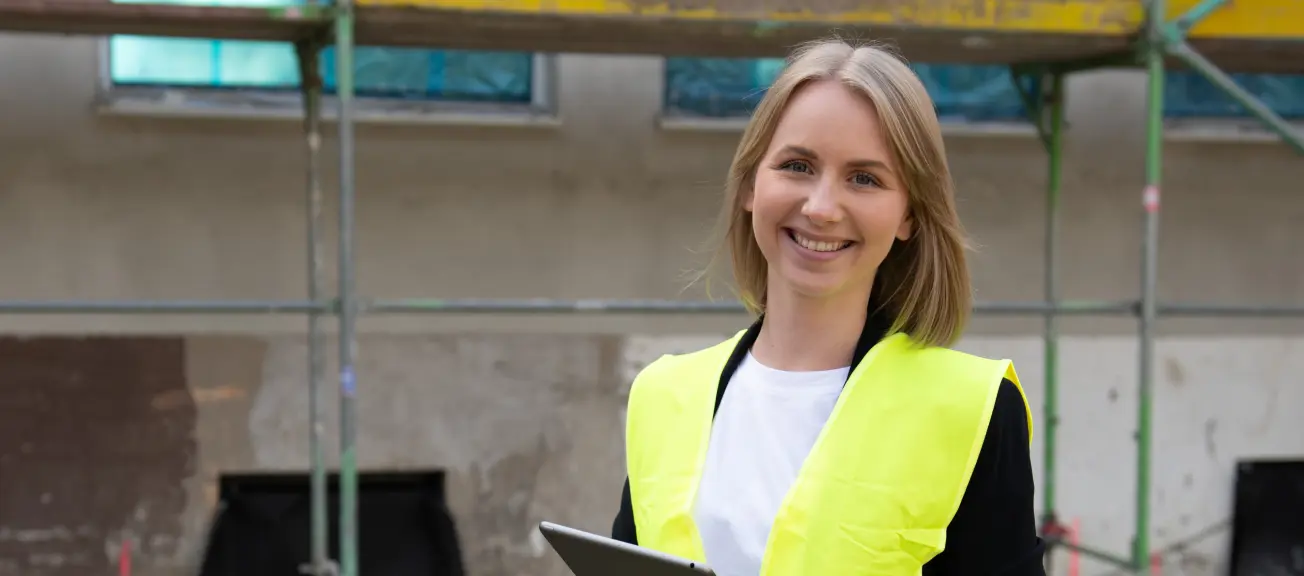 Eine lächelnde Frau, welche mit einer gelben Warnweste auf einer Baustelle steht und ein Ipad in der Hand hält