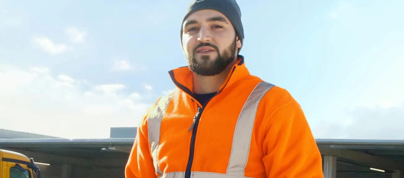 Ein junger Mann, welcher in Arbeitskleidung auf der Straße steht 