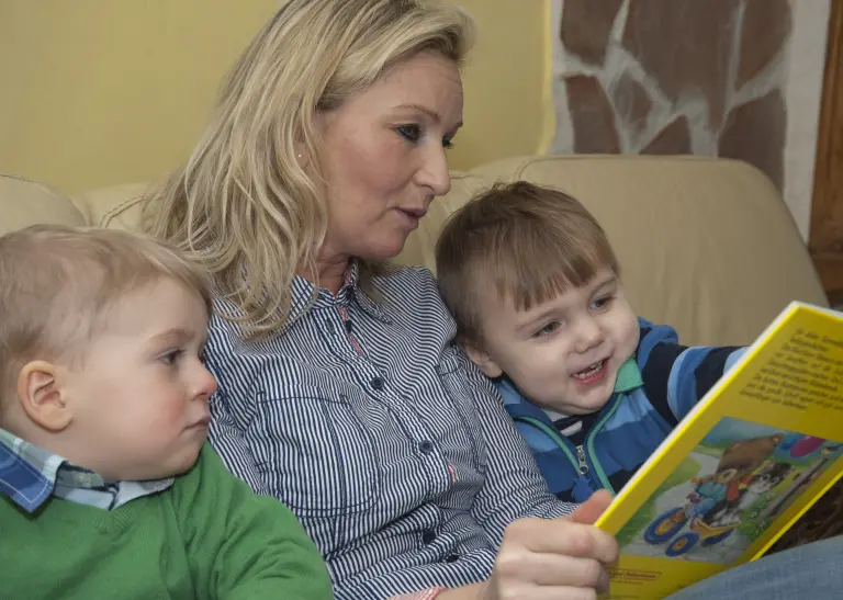 Eine blonde Frau sitzt zwischen zwei kleinen Jungen und liest ihnen ein großes gelbes Bilderbuch vor, während einer der Jungen mit seinem Finger auf eine Buchseite zeigt