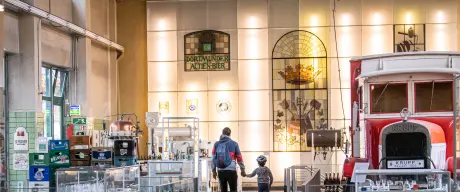 Ein Vater geht mit Kind an der Hand durch das Brauerei-Museum. Links und rechts stehen viele Biergläser, Bierkästen und Fässer.