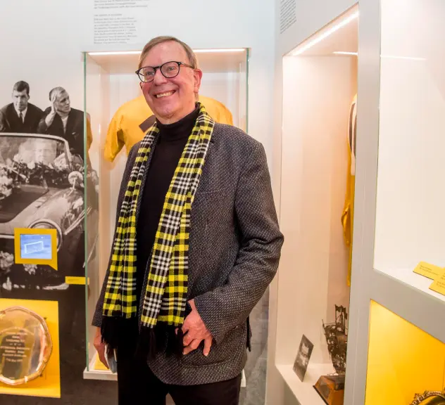 Zu sehen ist Gerd Kolbe in einem Ausstellungsraum voller BVB-Erinnerungsstücke. Er trägt einen schwarz-gelb karierten Schal und lacht in die Kamera.