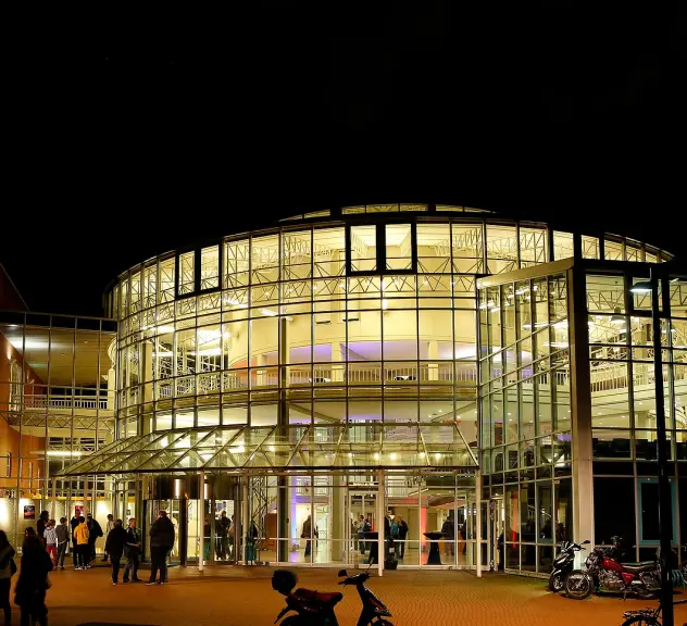 Außenansicht der DASA Arbeitswelt Ausstellung bei Nacht. Im zentrum des Bildes steht díe Glasfassade des Gebäudes, drinnen brennt Licht. Vor und in dem Gebäude stehen Menschen in Grüppchen. Links im Bild hängen an der Backstein-Außenwand drei Banner. Auf einem Banner steht "Mensch", auf einem steht "Arbeit" und auf dem dritten steht "Technik".