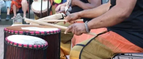 Mitglieder der Djolo Tanz Gruppe musizieren auf Trommeln. Im Hintergrund sitzt das Publikum auf einer Art kleinen Tribüne.