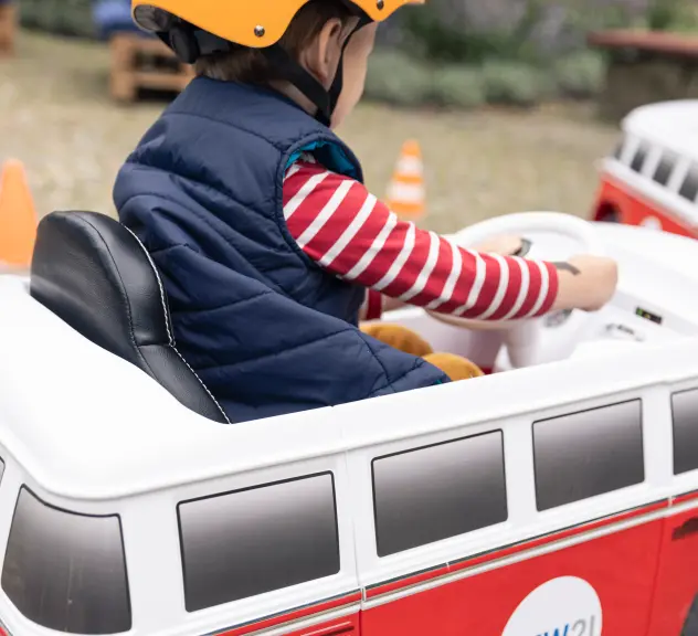 Zu sehen ist ein kleines Kind mit gelbem Helm in einem roten Miniatur-VW-E-Bus