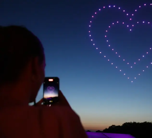 Zu sehen ist ein Nachthimmel, in dem eine Formation aus pink leuchtenden Drohnen schwebt. Die Drohnen bilden ein Herz. Im Vordergrund ist der Umriss einer Person zu sehen, die ein Bild der Formation mit ihrem Smartphone aufnimmt.