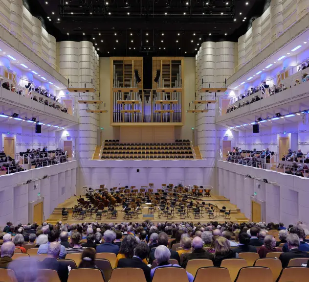 Zu sehen ist der große Saal des Konzerthauses. Mittig befindet sich die Fläche des Orchesters mit leeren Stühlen und einigen Instrumenten. Davor sowie links und rechtes an den Seiten befinden sich die Sitzplätze mit Zuschauer*innen.