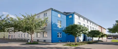 Zu sehen ist die Außenansicht des Künstlerhauses. Die Wände im mittleren Bereich sind blau, die anderen Wände weiß. Auf der blauen Wand steht außerdem „Künstlerhaus Dortmund“. Vor dem Haus ist ein Teil einer Straße zu sehen sowie ein paar Bäume.