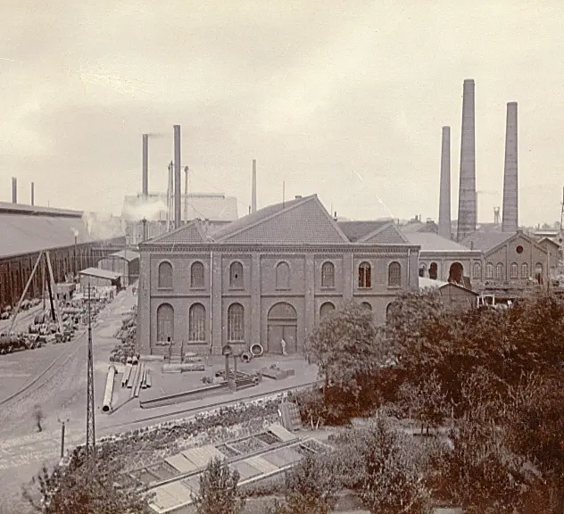 Blick auf das Hoesch-Werk. Im Vordergrund steht ein Fabrikgebäude aus Backstein. Vor dem Gebäude werden Gleise gebaut. Im Hintergrund sind viele rauchende Schlote zu sehen.