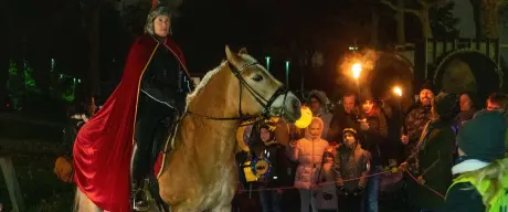 Zu sehen ist ein Mann auf einem Pferd verkleidet als Sankt Martin. Daneben sind Erwachsene und Kinder, die Laternen halten.