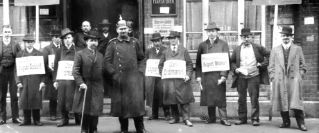 Eine Momentaufnahme zur Zeit des Reichtagswahlkampf 1912 in Dortmund. Verschiedene politische Anhänger stehen vor einem Lokal und tragen Wahlplakate um ihren Hals. Zwei Führungspersonen stehen vor den politischen Anhängern, alle Personen sind in Anzug gekleidet und die meisten tragen Hut.