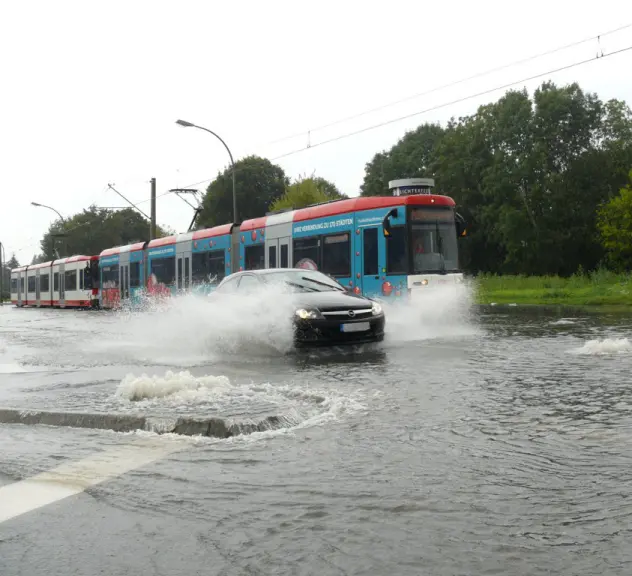 Ein dunkles Auto, das neben einer Stadtbahn über eine geflutete Straße fährt. Das Wasser spritzt und aus den Schachtdeckeln sprudelt das Abwasser.