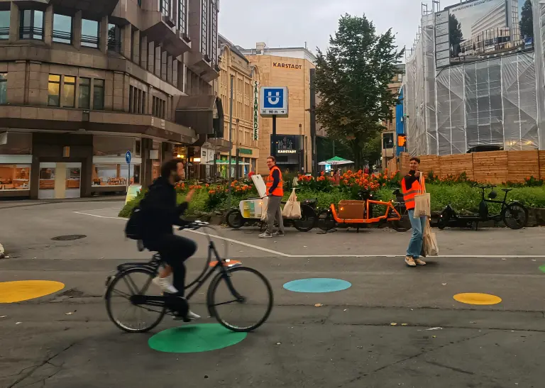 Zwei Männer in orangenen Warnwesten stehen auf der Kreuzung Kampstraße / Hansastraße. Sie halten Beutel in ihren Händen. Im Hintergrund stehen zwei Lastenräder. Ein Radfahrender fährt im Vordergrund von links nach rechts. Sein Gesicht ist unscharf, weil er in Bewegung ist. 