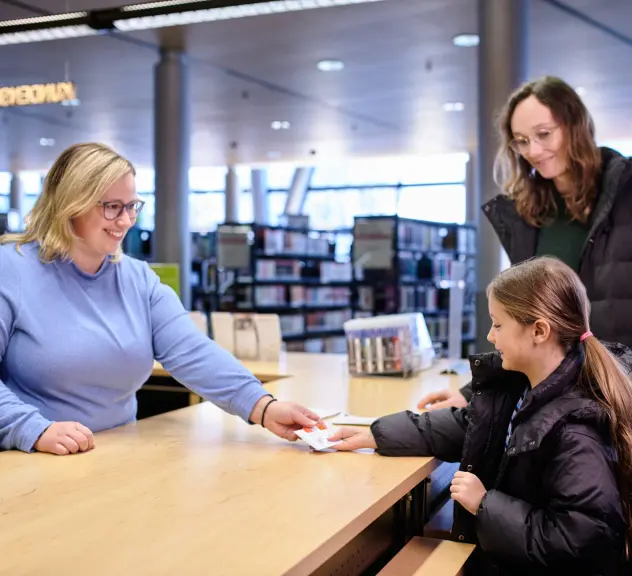 Bibliotheksmitarbeiterin überreicht einem Kind mit Mutter einen neuen Bibliotheksausweis