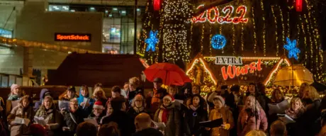 Viele Menschen stehen in der Weihnachtsstadt und singen. Im Hintergrund ist der Baum & bunte Lichter zu sehen