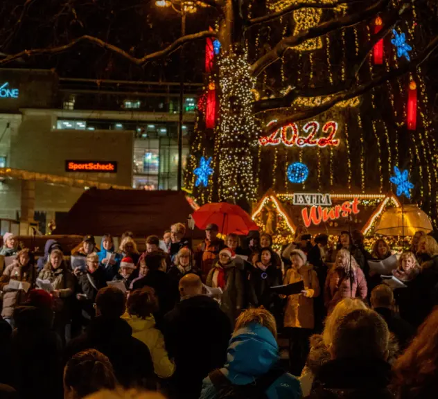 Viele Menschen stehen in der Weihnachtsstadt und singen. Im Hintergrund ist der Baum & bunte Lichter zu sehen