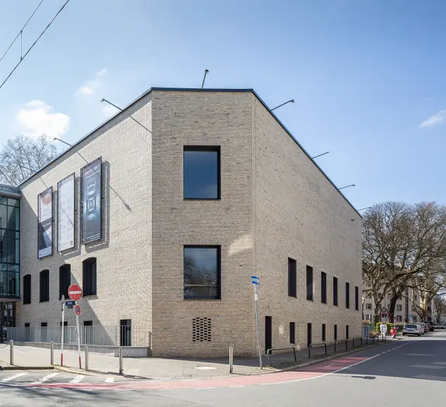 Ein modernes Gebäude mit heller Klinkerfassade, großen Fenstern und Werbebannern an der Seite, an einer Straßenecke gelegen.