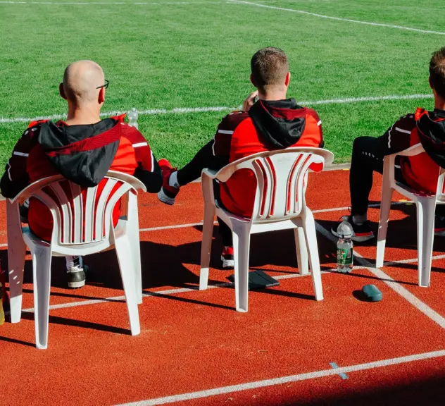 Fünf Personen sitzen mit dem Rücken zum Betrachter an einem Spielfeldrand