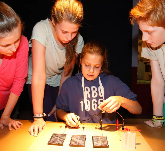 Drei Mädchen und ein Junge schauen sich ein elektrisches Mitmachexperiment mit einem Propeller im Kindermuseum auf einem Tisch an.