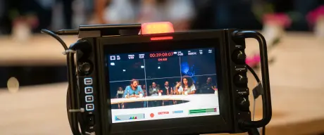 Ein Bild von einer Kamera in desse Fokus ein des Moderationsduo am Tisch ist.