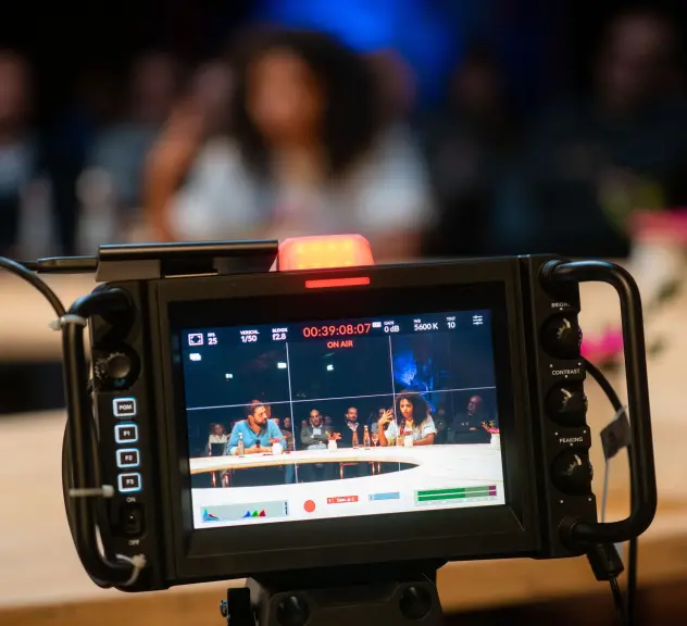 Ein Bild von einer Kamera in desse Fokus ein des Moderationsduo am Tisch ist.