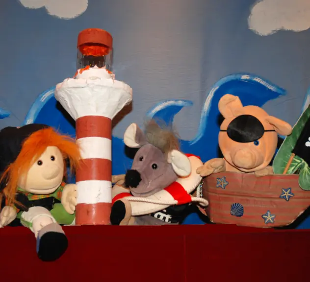 Eine Puppe, eine Maus und ein Schwein sind als Piraten verkleidet. In der Mitte steht ein Leuchtturm